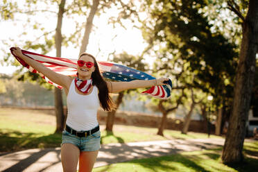 Porträt der patriotischen amerikanischen Frau, die mit der Nationalflagge im Park läuft und lächelt. Mädchen im Park mit USA-Flagge in den Händen, die den Unabhängigkeitstag feiert. - JLPSF19268