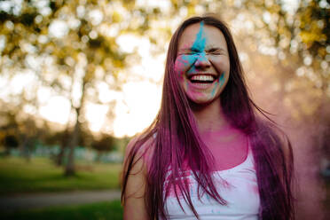 Porträt einer lächelnden jungen Frau, deren Gesicht mit Farben verschmiert ist. Fröhliches Mädchen im Park mit Farbpulver im Gesicht. - JLPSF19282