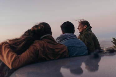 Eine Gruppe von Freunden steht auf der Autobahn und genießt die abendliche Aussicht von einem Hügel aus. Freunde stehen auf einer Höhenstraße mit ihrem Auto und genießen die Abenddämmerung. - JLPSF19292