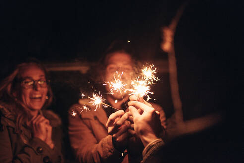 Group of friends lighting fire sparkle sticks in the dark and enjoying. Man holding burning fire sparkle sticks in hands enjoying with friends. - JLPSF19297