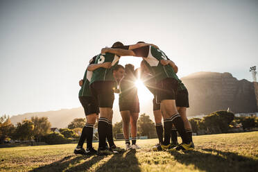 Rugby players standing in a circle with their hands on shoulders. Rugby team in huddle after the match. Bright sunshine through the huddle. - JLPSF19310