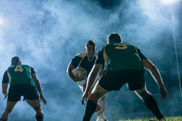 Ein professioneller Rugbyspieler läuft mit einem Ball und gegnerischen Spielern, die versuchen, ihn während eines Nachtspiels in einer Sportarena zu blockieren. - JLPSF19320