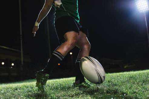 Rugby-Spieler lässt den Ball auf den Boden fallen und kickt ihn dann, während er aufspringt. Rugby-Spieler, der unter den Lichtern einer Sportarena ein fallengelassenes Tor trifft. - JLPSF19330
