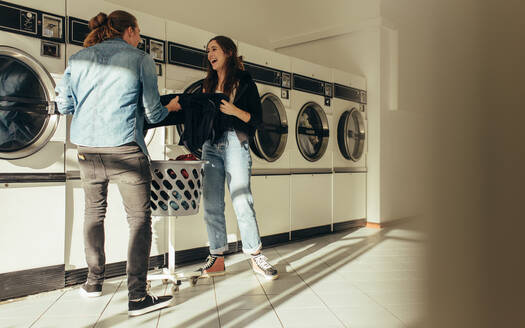 Ein lächelnder Mann und eine lächelnde Frau, die zusammen Wäsche in eine Waschmaschine stecken, stehen in einer Waschküche. Ein glückliches Paar, das zusammen Wäsche wäscht. - JLPSF19337