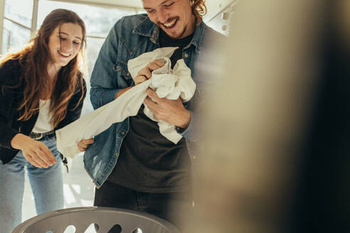 Smiling man and woman putting clothes in a washing machine together. Happy couple having fun doing laundry together. - JLPSF19339