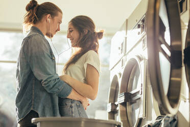 Mann und Frau hören Musik und teilen sich Kopfhörer, die zusammen in einer Waschküche stehen. Intimes Paar, das sich gegenseitig in einer Waschküche hält. - JLPSF19350