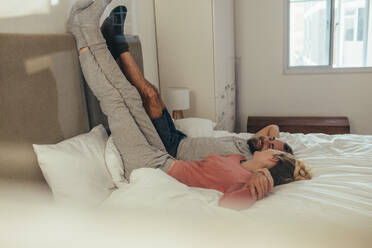 Ein Paar liegt zu Hause auf dem Bett, hat die Beine an die Wand gelehnt und unterhält sich. Ein glücklicher Mann und eine glückliche Frau liegen auf dem Bett und halten sich an den Händen. - JLPSF19356