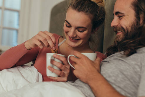 Lächelnder Mann und Frau mit ihren Kaffeetassen auf dem Bett sitzend. Paar sitzt auf dem Bett unter einer gemütlichen Decke und genießt den Kaffee zusammen. - JLPSF19357
