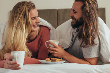 Happy couple holding coffee cups relaxing on bed with snacks. Smiling couple lying on bed in a cozy blanket looking at each other while having breakfast. - JLPSF19358