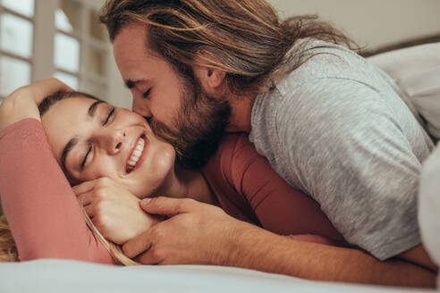 Paar im Bett verbringt Zeit zusammen mit Mann küsst seine Partnerin. Lächelnde Frau mit geschlossenen Augen wird geküsst und geliebt von ihrem Mann. - JLPSF19362