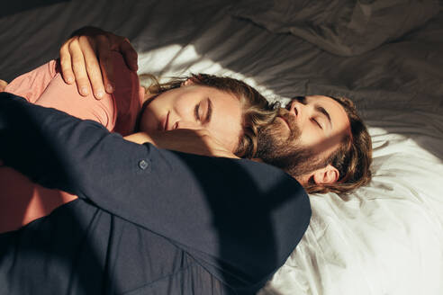 Frau schläft auf dem Arm ihres Mannes im Bett mit Sonnenlicht, das aus dem Fenster fällt. - JLPSF19366