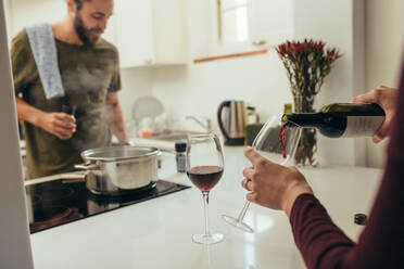 Mann kocht Essen in der Küche, während Frau Wein in Gläser gießt. Paar in der Küche bereitet sich auf das Abendessen mit Essen und Wein vor. - JLPSF19370