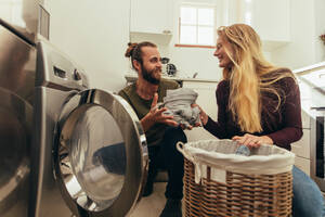 Happy couple putting clothes in washing machine. Couple using a front loading washing machine to wash laundry. - JLPSF19376