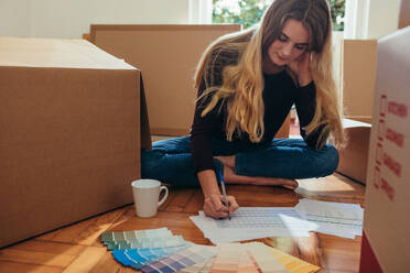Eine Frau sitzt auf dem Boden, um sich herum Umzugskartons, und erstellt eine Checkliste. Eine Frau wählt anhand von Farbkarten eine Wandfarbe für ihr neues Haus aus. - JLPSF19382