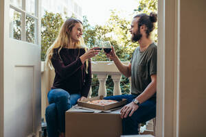 Ein Paar sitzt in seinem neuen Haus, isst Pizza und trinkt Wein. Ein Paar feiert seinen Einzug in ein neues Haus mit Wein und Pizza. - JLPSF19388