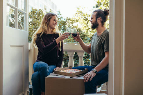 Ein Paar sitzt in seinem neuen Haus, isst Pizza und trinkt Wein. Ein Paar feiert seinen Einzug in ein neues Haus mit Wein und Pizza. - JLPSF19388