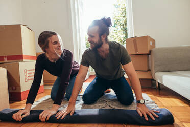 Smiling man and woman sitting on a carpet and rolling it for packing. Couple looking at each other while packing their household items. - JLPSF19390