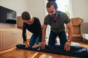 Smiling man and woman packing their household items in packing boxes. Couple working together to roll a carpet on floor for packing. - JLPSF19391