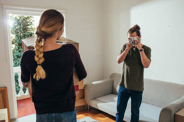 Mann, der eine Frau fotografiert, die einen Umzugskarton in ein neues Haus trägt. Rückansicht einer Frau, die einen Umzugskarton trägt, während ihr Partner sie mit einer Digitalkamera fotografiert. - JLPSF19392