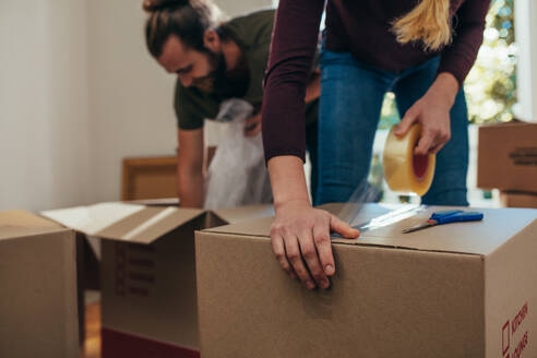 Couple using packing boxes to pack their household items. Man putting items in boxes while a woman applies adhesive tape on the boxes to seal them. - JLPSF19396