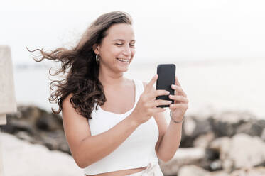 Lächelnde Frau, die am Wochenende am Strand mit ihrem Smartphone fotografiert - MEF00125