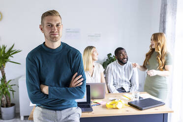 Confident businessman with arms crossed by colleagues in background at office - WPEF06665