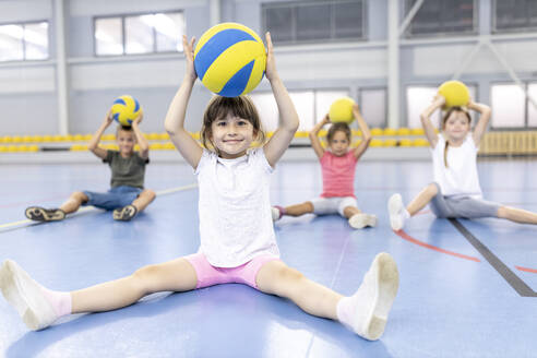 Lächelndes Mädchen, das einen Ball über dem Kopf hält und mit Freunden auf dem Schulsportplatz sitzt - WESTF25145