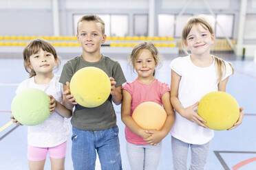Smiling elementary students with ball standing side by side at school sports court - WESTF25150