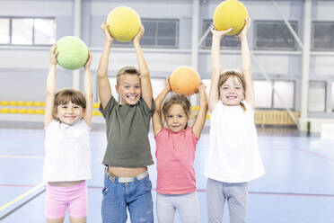 Cheerful students with arms raised holding ball standing together at school sports court - WESTF25151