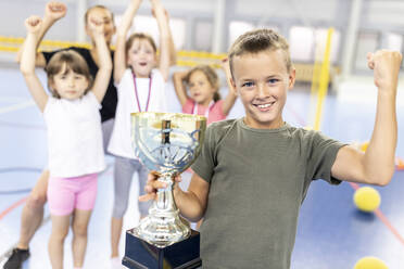 Happy boy flexing muscle holding trophy with friends at school sports court - WESTF25158