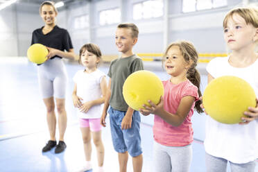 Smiling students holding ball with teacher standing at school sports court - WESTF25175