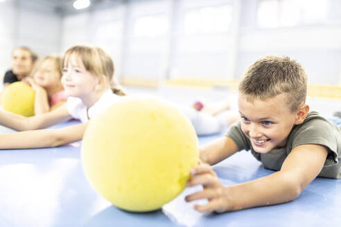 Lächelnder Junge im Grundschulalter betrachtet einen Ball, der auf dem Schulsportplatz liegt - WESTF25179
