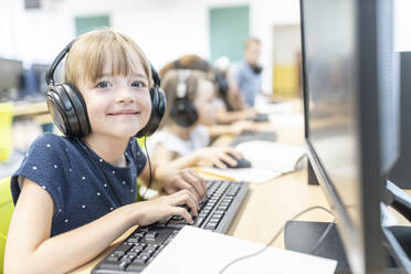 Lächelndes Mädchen mit Pony, das kabelgebundene Kopfhörer trägt und im Computerkurs in der Schule sitzt - WESTF25203