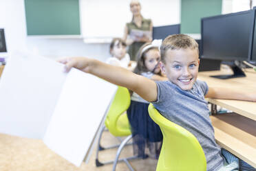 Aufgeregter Junge mit Notebook auf einem Stuhl im Klassenzimmer sitzend - WESTF25221