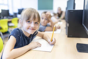 Lächelndes Mädchen mit Bleistift und Buch sitzt am Schreibtisch im Klassenzimmer - WESTF25227