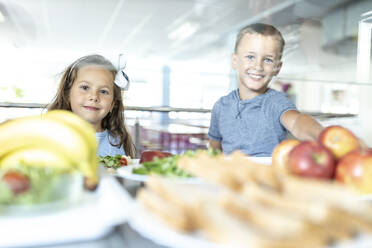 Lächelnde Grundschüler, die in der Mittagspause in der Schulcafeteria Obst essen - WESTF25244