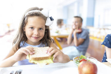 Smiling girl holding sandwich in school cafeteria - WESTF25256