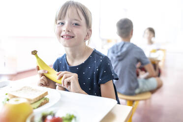 Lächelnder Schüler mit frischer Banane in der Mittagspause in der Cafeteria - WESTF25258