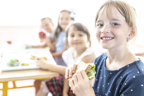 Student hält frisches Sandwich mit Freunden im Hintergrund in der Cafeteria - WESTF25263