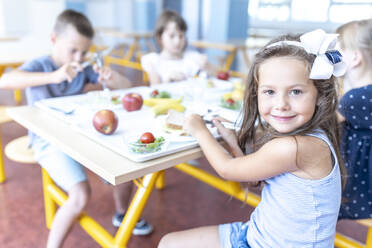 Lächelndes Mädchen beim Mittagessen mit Freunden, die am Tisch in der Schulcafeteria sitzen - WESTF25266