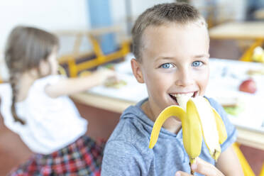 Glücklicher Junge mit blauen Augen isst Banane in der Mittagspause in der Cafeteria - WESTF25270