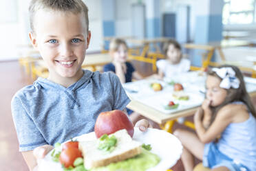Smiling boy holding healthy lunch on plate standing at cafeteria - WESTF25273