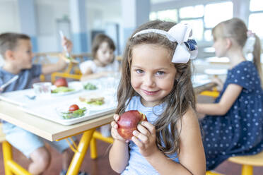 Lächelndes Mädchen mit Apfel in der Hand sitzt mit Freunden in der Schulcafeteria - WESTF25275