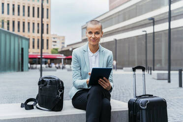 Lächelnde Geschäftsfrau mit Tablet-PC und Gepäck auf einer Bank am Fußweg sitzend - OIPF02522