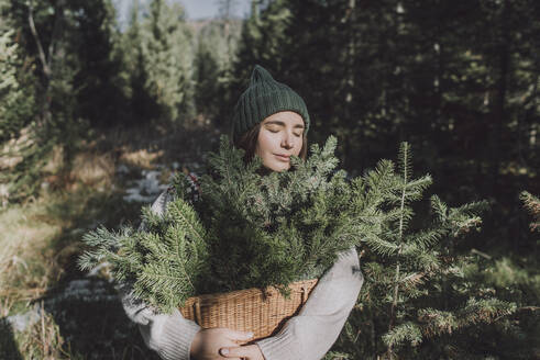 Woman with eyes closed holding basket of spruce twigs at forest - VBUF00194