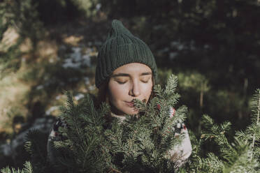 Frau mit geschlossenen Augen riecht an den Zweigen eines immergrünen Baumes - VBUF00197