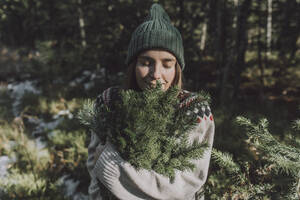 Woman with eyes closed smelling twigs of evergreen tree in forest - VBUF00198