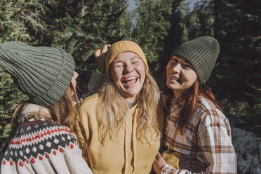 Frauen halten Weihnachtsbaumzweige hinter der Strickmütze ihrer Freundin im Wald - VBUF00206