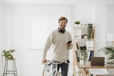 Smiling businessman with bicycle and smart phone in office - EBBF06734