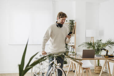 Businessman with bicycle text messaging on smart phone in workplace - EBBF06735